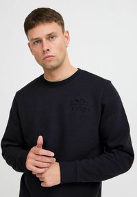 Zwarte sweatshirt gemaakt van een katoenmix, met een gestructureerd logo op de borst en geribbelde manchetten. De handen van het model zijn samengevouwen.