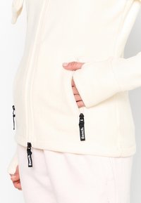 Giacca in fleece crema con tasche con zip; presenta dettagli in metallo nero e un design con foro per il pollice sui polsini per calore e comfort.