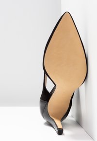 Chaussure à talon aiguilles noire, positionnée semelle vers le haut, montrant une semelle beige lisse et un bout pointu sur un fond blanc avec coins.