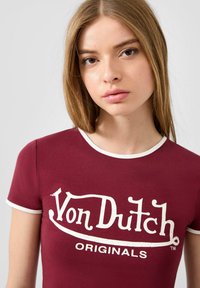 Bordeaux gekleed katoenen t-shirt met een witte ribbelkraag, met een verouderd logo in het wit met "Von Dutch" en "ORIGINALS."