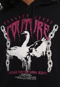 Black hoodie featuring an oversized white swan graphic adorned with rhinestones, pink lettering, and chain accents.