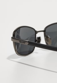 Sorte solbriller med mørke glas. Stellet har et slankt, rundt design med "QUAY"-logoet på stængerne. Glansfuld finish.