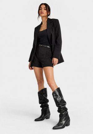 Frau steht und trägt einen schwarzen Blazer, ein schwarzes Oberteil, schwarze Shorts mit Gürtel und kniehohe schwarze Lederstiefel vor einem schlichten weißen Hintergrund.