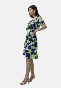 Vestido floral en verde con grandes flores azul marino, mangas cortas abullonadas, detalles de fruncido y un dobladillo a la altura de la rodilla. Complementado con tacones con tiras.
