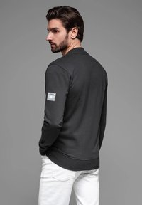 Sudadera gris oscuro de manga larga, con cuello redondo y una etiqueta de la marca en el brazo izquierdo. Combinada con pantalones blancos, que muestran un detalle de dobladillo acanalado.