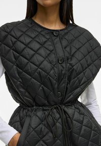 Quiltad svart väst med hög krage, med svarta knappar, elastisk dragsko i midjan och en slät, lätt glänsande yta.