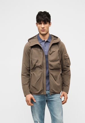 Νεαρός άνδρας με καφέ μπουφάν με κουκούλα πάνω από μπλε πόλο και ανοιχτόχρωμα σκισμένα τζιν παντελόνια, στέκεται μπροστά σε απλό φωτεινό φόντο.