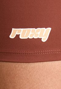 Brune atletisk shorts med en glatt tekstur har en hvit logo "roxy" i en stilisert font langs livlinningen.