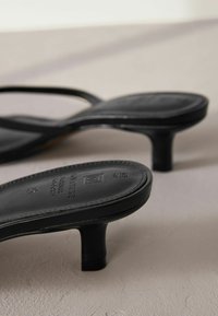 Sandali neri con tacco alto, dotati di cinturini sottili e una texture in pelle liscia. Presentano un tacco cilindrico basso e un marchio impresso sulla suola.