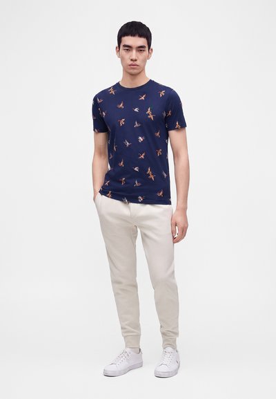 Νέος άνδρας στέκεται φορώντας μπλε μπλουζάκι με σχέδια πουλιών, μπεζ παντελόνια και λευκά αθλητικά σε ένα απλό φωτεινό φόντο.