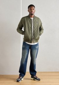 Veste bomber matelassée vert olive avec des poignets en côtelé, associée à un jean droit bleu délavé et à des baskets multicolores.