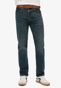 Jeans de mezclilla de color azul oscuro con corte recto, que presentan un cinturón marrón. Los jeans tienen un lavado sutil y un diseño clásico de cinco bolsillos.
