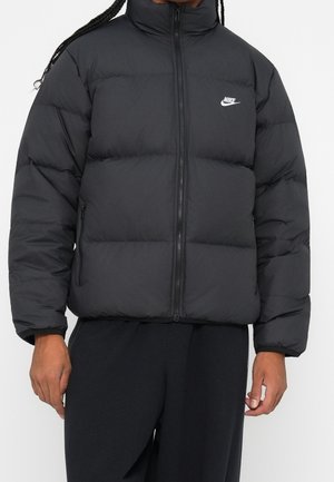 Black quilted puffer jacket with front zipper and white Nike logo on chest, worn by a person with dark pants against a plain background.