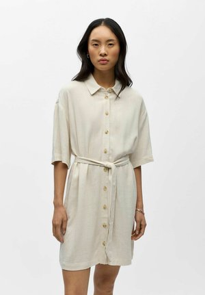 Mujer de pie, vestida con un vestido beige hasta la rodilla, de manga corta y con botones, con un cinturón atado en la cintura, sobre un fondo blanco liso.