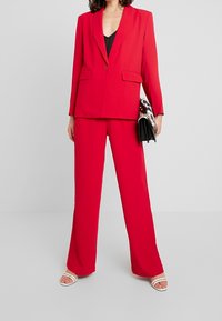 Roter, maßgeschneiderter Anzug bestehend aus einem taillierten Blazer mit einem Knopf und passenden weit geschnittenen Hosen. Accessoires sind eine schwarze Clutch.