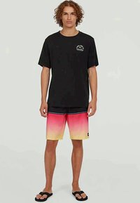 Camisa preta de manga curta com logotipo, acompanhada de calções de banho em degradê que vão do rosa ao amarelo. Sandálias planas.