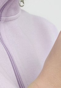 Prenda de color púrpura claro con cremallera y cuello alto, que presenta un tirador de cremallera plateado y una tela texturizada. Primer plano de la zona de la cremallera.