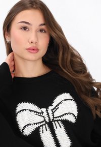 Pulover negru cu un design mare de fundă albă dintr-un material texturat, cu accente de cusături. Modelul are părul lung, castaniu și cercei transparenți.