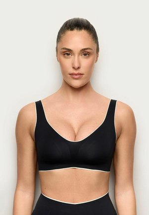 Mujer con el cabello recogido, vistiendo un sujetador deportivo negro y pantalones de talle alto, sobre un fondo claro y liso.