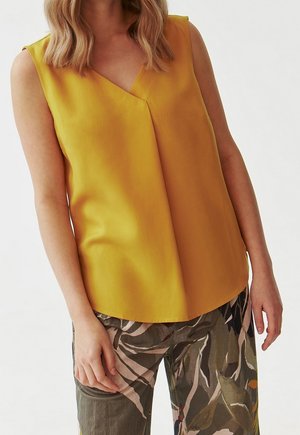 Kvinde iført ærmeløs sennepsgul V-hals top og løse mønstrede bukser med abstrakte bladmotiver.