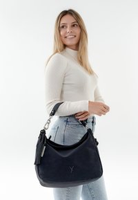 Svart hobo-väska tillverkad av texturerad tyg med en slät yta, med ett handtag i silverfärgad metall och en dekorativ tofs.