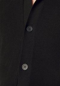 Gros plan sur un cardigan en maille côtelée noire avec deux boutons noirs, chacun gravé du nom de la marque "Sandro".