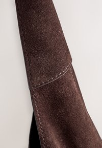 Gros plan d'une sangle en daim marron avec des coutures visibles et une surface douce et texturée sur un fond clair.