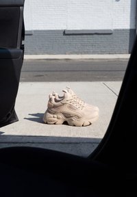 Paire de baskets beiges épaisses posées sur un trottoir ensoleillé à côté d'une porte de voiture ouverte avec un mur en briques gris et blanc en arrière-plan.