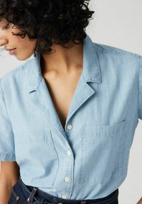 Light blue short-sleeve button-up shirt made of denim, featuring a collar and two front pockets. Classic casual design with a relaxed fit.