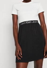 Šaty s krátkými rukávy s bílým žebrovaným topem a černou, splývavou sukní. Mají široký černý pás s nápisem "Calvin Klein Jeans" bílým písmem.