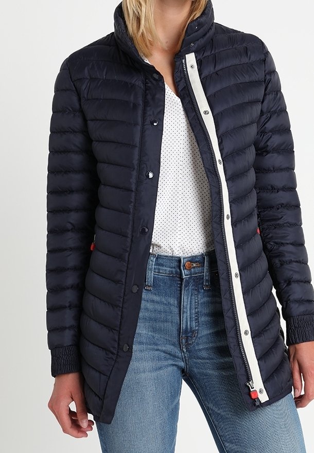 Marineblauwe donsjas met doorgestikt ontwerp, opstaande kraag, witte ritssluiting aan de voorkant en rode accenten. Draagt een wit shirt met stippen en een spijkerbroek eronder.