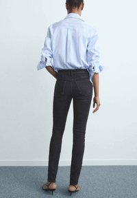 Persona de pie de espaldas a la cámara, vistiendo jeans negros ajustados, una camisa de botones azul claro con mangas enrolladas y tacones negros con tiras.
