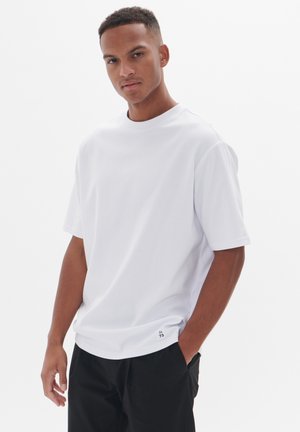 Kurzärmliges, rundhalsiges, weißes T-Shirt aus glattem Stoff, mit einem gestickten Logodetail am Saum, kombiniert mit schwarzen Hosen.