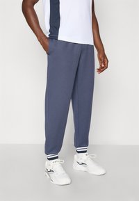 Μπλέ σκούρες φόρμες με πλαϊνές τσέπες, ριμπ μανσέτες στους αστραγάλους με λευκές και μπλέ ρίγες, συνδυασμένες με λευκά αθλητικά παπούτσια.