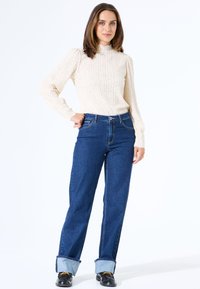 Crèmekleurige sweater van textuur met hoge hals, gecombineerd met blauwe wijde jeans met opgerolde mouwen en zwarte veterschoenen. Witte achtergrond.