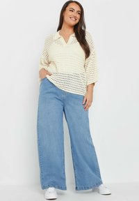 Krämfärgad virkad topp med V-ringning och krage, kombinerad med vida ljusblå denimjeans. Modellen bär vita sneakers.
