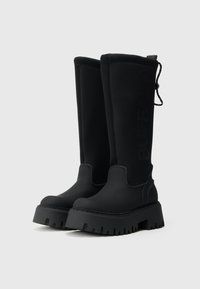 Melnas ceļgala augstuma zābakus ar masīvu zoli, kas izgatavoti no gluda gumijai līdzīga materiāla. Iekļauta izcelta 'BUFFALO' zīmola emblēma un regulējams aukliņu stiprinājums.