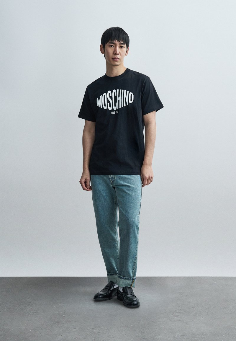 T-shirt en coton noir avec le logo « MOSCHINO », associé à un jean en denim bleu clair. La tenue est complétée par des mocassins noirs. Fond minimaliste.
