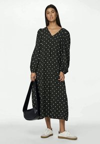 Μαύρο μακρύ φόρεμα με λευκό σχέδιο κουκίδες, V λαιμόκοψη και φουσκωτά μανίκια. Το μοντέλο κρατάει μια μαύρη τσάντα και φοράει λευκά αθλητικά παπούτσια με καφέ σόλες.