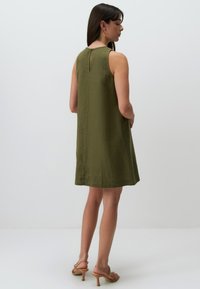 Olivenfarbendes ärmelloses Kleid mit glatter Textur, knielangem Schnitt und einem Rückenschlitz-Detail. Kombiniert mit hellbraunen Riemchen-Heels.