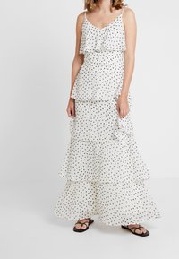 Vestido maxi blanco en capas con lunares negros, con capas con volantes y tirantes finos. Estilizado con sandalias planas negras.