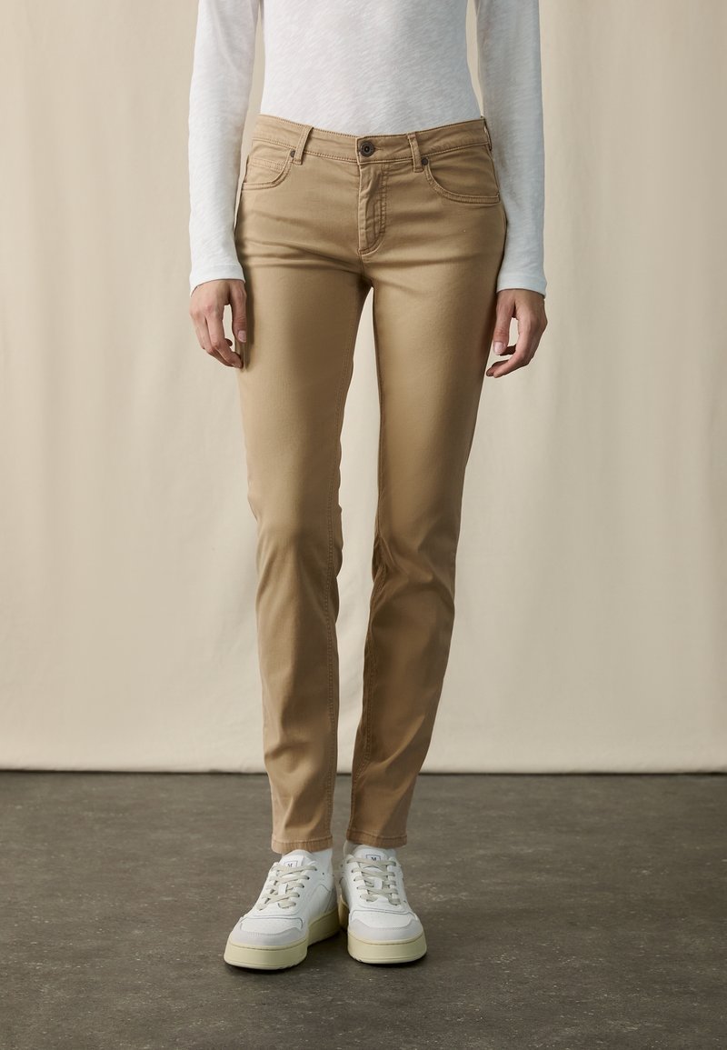Personne portant un pantalon beige ajusté, une chemise blanche à manches longues et des baskets blanches, debout sur un sol gris devant un fond beige.