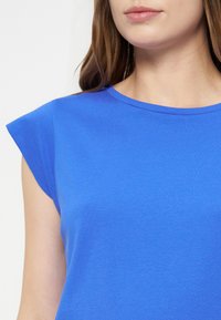 Femme portant un haut bleu vif à manches courtes et col rond, montrant le haut du torse et une partie de son visage et de ses cheveux.