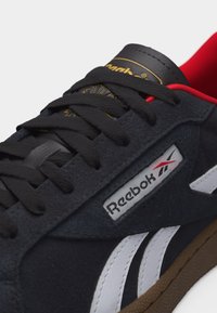 Zapatilla negra de Reebok con cordones negros, rayas laterales blancas, forro interior rojo y suela color goma, mostrando el logo de la marca y la etiqueta de la lengüeta.