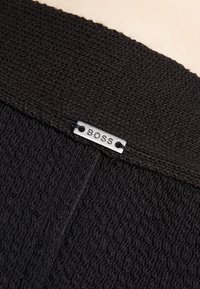 Černá pletená tkanina s texturovaným designem, na horním okraji je připevněna malá kovová značka s vyrytým nápisem "BOSS".