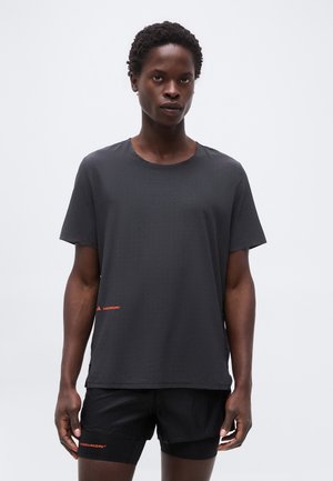 Férfi fekete rövid ujjú, perforált sportpólóban és fekete rövidnadrágban, mindkét ruhadarabon kis piros logós felirattal, fehér háttér előtt áll.