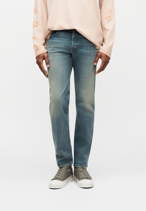 Inimene, kes kannab helesiniseid teksapükse, beeži pika varrukaga särki ja hallid kõrge säärega tossud, seisab valgel taustal.