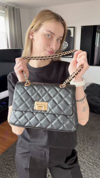 Joven con cabello claro sostiene un bolso negro acolchado con cadena dorada y cierre en una sala de estar moderna.