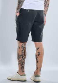 Dark fabric shorts with a simple design, side pockets, and a button detail. Worn with light-colored sneakers and visible tattoos on the legs.
