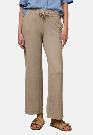 Frau trägt beige strukturierte Hose mit Kordelzug, blaues kurzärmeliges Hemd und braune offene Slip-on-Sandalen mit Schnallendetails.